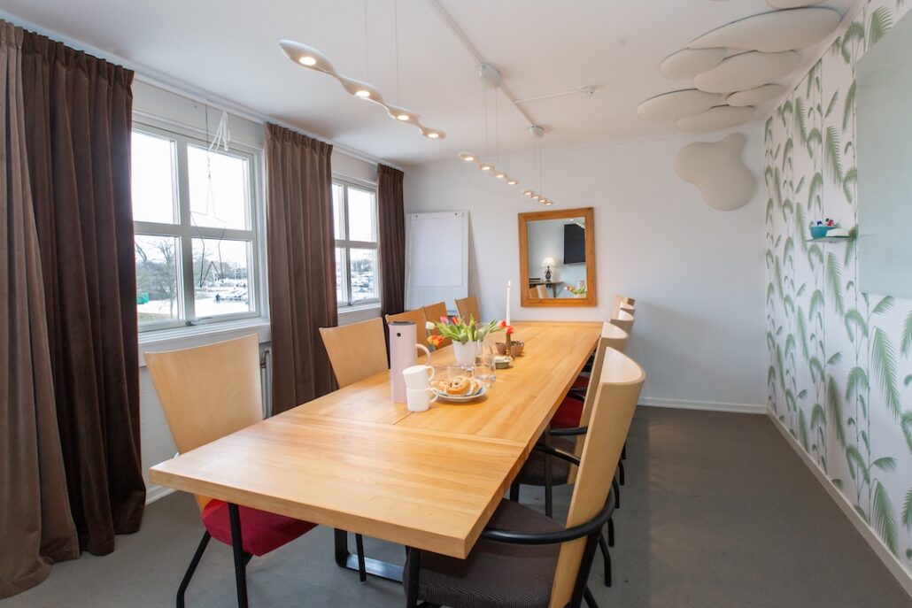 Ljust och välorganiserat konferensrum under Almedalsveckan med ett stort träbord, bekväma stolar och modern belysning, inrett med ett fönster som vetter mot visby ringmur och en vägg dekorerad med ett tropiskt bladmönster.