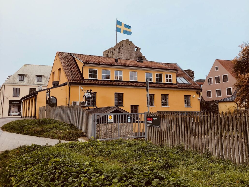 Vy av 'Kontoret' vid Österport, beläget i ett iögonfallande gult hus i Visby centrum. Huset är omgivet av den historiska ringmuren med synliga stenstrukturer. Ett svenskt flagga vajar ovanför byggnaden, och en cykel är parkerad vid staketet i förgrunden