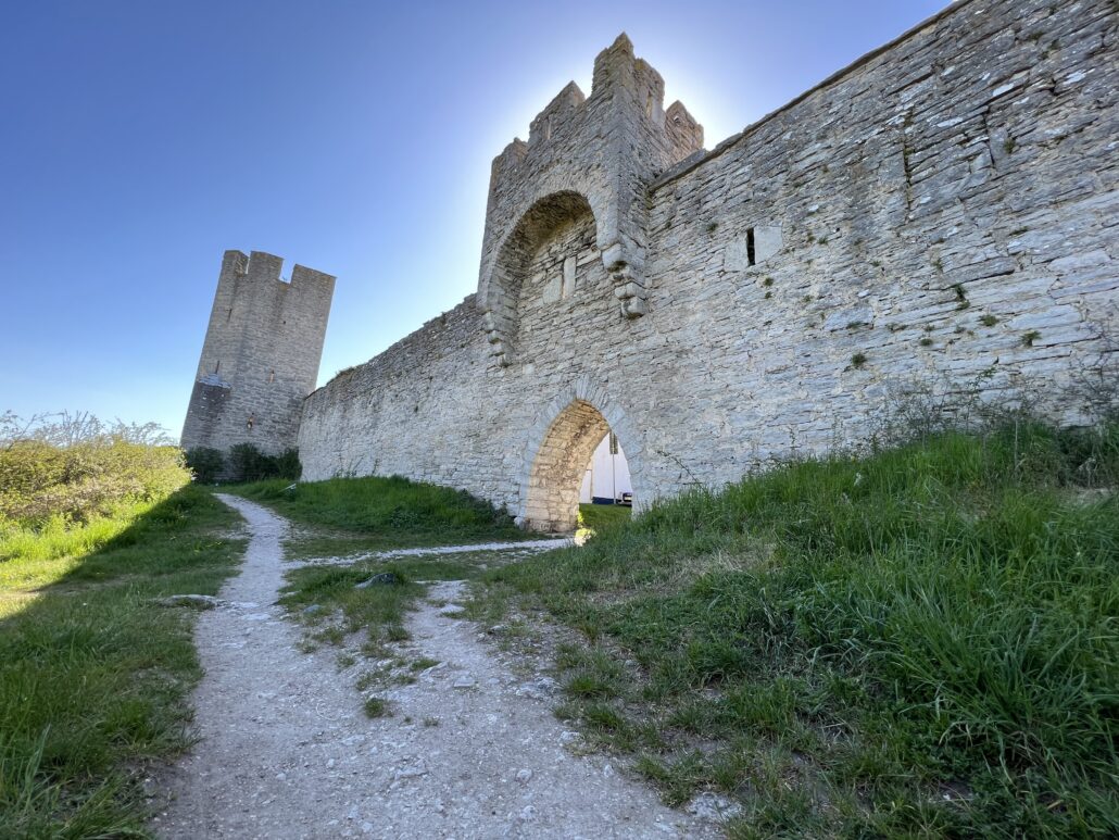 Stig av grus som leder fram längs utsidan av Visby ringmur en solig dag. Ett högt medeltida torn lutar svagt mot vänster, och ett valv i muren släpper in solljus genom öppningen. Grönt gräs växer längs båda sidor av stigen, och himlen är klarblå utan moln.