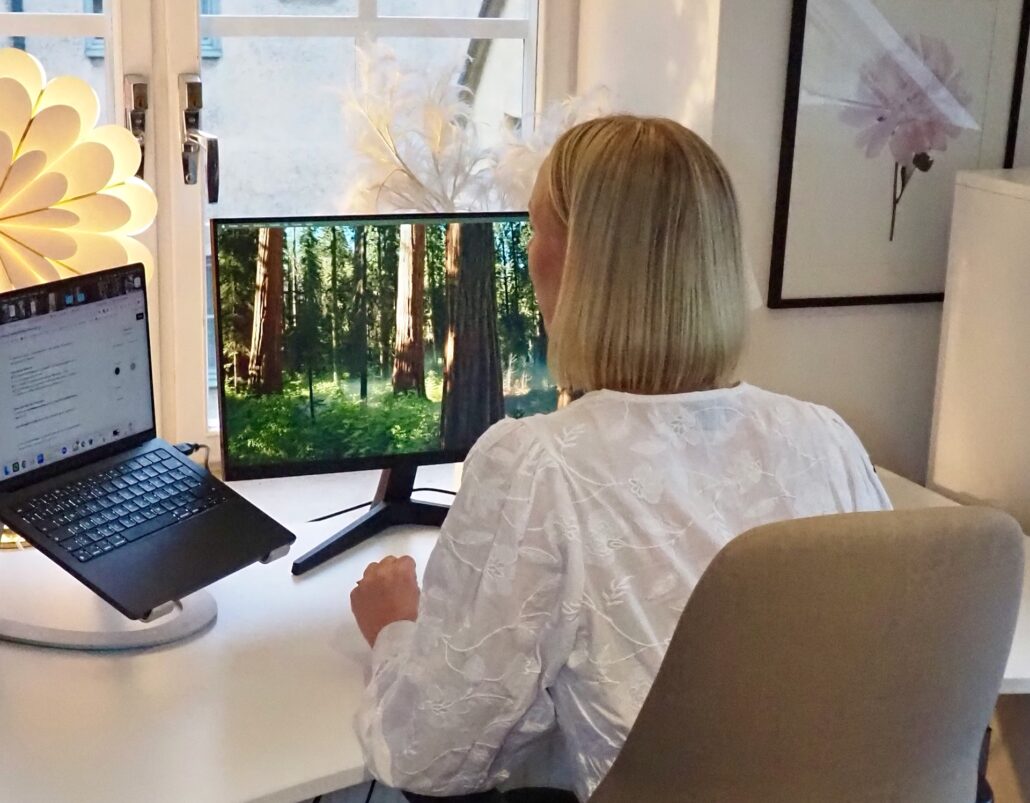 Linnéa sitter vid sitt skrivbord på Kontoret vid Österport i Visby och arbetar framför två skärmar, där den ena visar en bild av en solbelyst skog. Ljuset från fönstret och den moderna inredningen skapar en lugn och inspirerande arbetsmiljö.